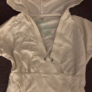 Vit hoodie med kort ärm från H&M Sport - Säljer en vit hoodie från H&M Sport i storlek M. Tröjan har korta ärmar, djup v-ringning med snörning och en stor huva. Den säljs inte längre och jag fått ärva den av min mamma då hon bärde den i 2000s talet så perfekt för dig som gillar y2k/oldschool. Väldigt fin och perfekt för att ha t.ex ett linne under.