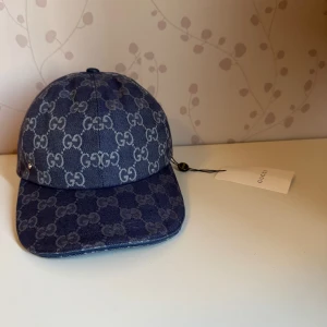Blå keps från Gucci med monogram - Säljer en blå keps från Gucci i jeansmaterial med klassiskt GG-monogram i ljusgrått över hela kepsen. Kepsen har böjd skärm och justerbar rem baktill. Perfekt för dig som gillar exklusiva accessoarer.