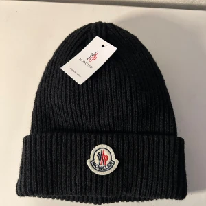 Sprillans ny! Moncler mössa - Svart ribbstickad mössa från Moncler med uppvikt kant och klassisk Moncler-logga framtill. Perfekt accessoar för kyliga dagar och enkel att matcha med olika outfits.