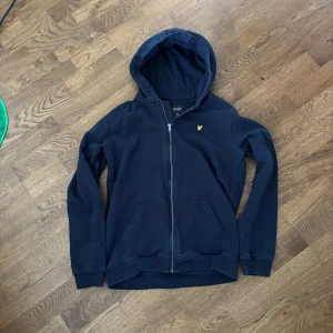 Mörkblå hoodie från Lyle & Scott - Säljer en mörkblå hoodie från Lyle & Scott med dragkedja och huva. Tröjan har två fickor framtill och en gul broderad logga på bröstet. Perfekt för en avslappnad stil. 