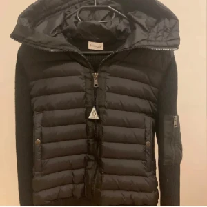 Moncler cardigan - Säljer en svart pufferjacka från Moncler med stickade ärmar och huva. Jackan har dragkedja framtill, två fickor med knappar och en extra ficka på ärmen. Den är i storlek L men passar M