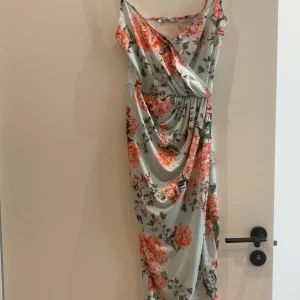 Blommig långklänning från NLY One - Säljer en superfin långklänning från NLY One med blommigt mönster i rosa och grönt på ljus bakgrund. Klänningen har omlottliv, smala axelband och markerad midja. Perfekt för sommarens alla tillfällen!