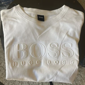 Vit långärmad tröja från Hugo Boss - Säljer en stilren vit långärmad tröja från Hugo Boss med upphöjd logotyp framtill. Tröjan har rund halsringning och är tillverkad i mjukt material. Perfekt för en clean och klassisk look.