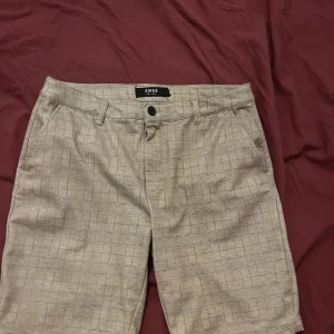Beige rutiga shorts från SMOG, aldrig använts - Snygga beige shorts från SMOG med diskret rutigt mönster. Klassisk passform med fickor fram och bak samt bälteshällor. Perfekta för varma dagar när du vill vara både stilren och bekväm.