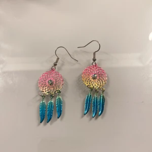 Färgglada drömfångar-örhängen med fjädrar - Säljer ett par örhängen i form av drömfångare med metallfjädrar. Örhängena har en gradient i rosa, gult och blått samt en liten diamant i mitten. Perfekta för att ge en bohemisk touch till din stil. Köpta från Glitter. 💖 Medföljer ploppar, jag rengör alltid örhängena innan jag skickar!! 