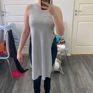 Grå ärmlösa T-shirts från H&M Divided - Säljer en grå ärmlös lång t-shirts från H&M Divided. T- shirts är ribbad och har höga slitsar på sidorna vilket ger en cool och modern look. Perfekt att styla med jeans eller leggings för en avslappnad vibe.