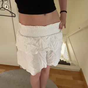 Vit volangkjol med smock i midjan - Säljer en söt vit kjol med bred smock i midjan och dubbla volanger. Kjolen är kort och har en luftig känsla, perfekt för varma dagar. Passar dig som gillar en feminin och trendig stil.