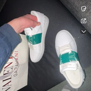 Valentino sneakers med grön detalj - ANVÄNDIT ENDAST 1 GÅNG, NI VET NOG VARFÖR DOM ÄR SÅ BILLIGA. Säljer ett par vita Valentino sneakers med en bred grön rem över vristen och gulddetaljer på tungan. Skorna har platt sula och rund tå, samt klassisk Valentino-logga. Perfekta för dig som vill sticka ut med stilrena men unika sneakers.