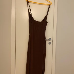Brun långklänning med slits och tunna axelband - Säljer en brun långklänning med tunna axelband och draperad urringning. Klänningen har en slits framtill och är figurnära i modellen. Perfekt för dig som gillar en stilren och elegant look.