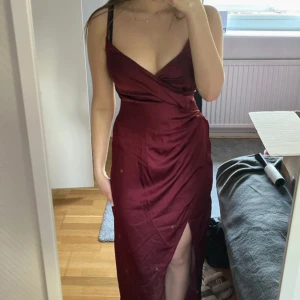 Vinröd långklänning med slits och omlott - Säljer en vinröd långklänning med smala axelband och omlottdesign framtill. Klänningen har en snygg slits och v-ringad urringning, perfekt bal. Hade själv denna på balen i högstadiet, vilket är enda gången den blivit använd. Den högra kupan är väldigt stor Förstod själv inte hur den skulle se ut Men jag tog säkerhetsnålar När jag använde den för att få det att se ut som hur jag försöker visa på andra bilden, blir superfin detalj! 💗 lite skrynklig men går ju såklart att fixa🥰