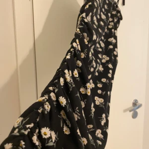 Svart blommig långklänning med smala axelband - Säljer en svart långklänning med vita och gula blommor. Klänningen har smala axelband och en åtsittande passform med rynkade detaljer längs sidan. Perfekt för sommarens tillfällen!