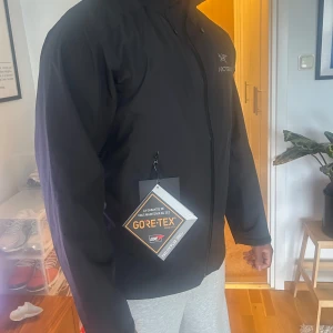 Svart vindjacka från Arc'teryx med GORE-TEX - Svart vindjacka från Arc'teryx med GORE-TEX-material. Jackan har dragkedja framtill och huva. Perfekt för utomhusaktiviteter och skyddar mot regn och vind. Enkel och stilren design med diskret logga på bröstet.