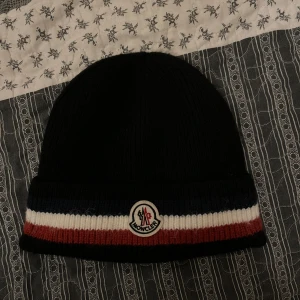 Moncler mössa - Svart ribbstickad mössa från Moncler med bred uppvikt kant i blått, vitt och rött. Klassisk Moncler-logga framtill. Perfekt för kyliga dagar. Pris kan diskuteras och byten funkar