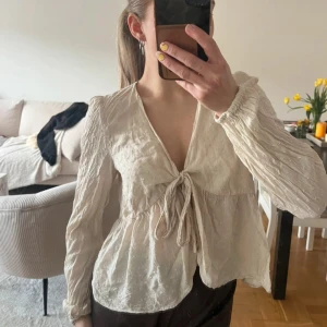 Krämvit blus med knyt från VILA - Säljer en krämvit blus från VILA med lång ärm och knyt framtill. Blusen har lätt struktur i tyget och v-ringning. Perfekt för vår och sommar, och passar till både jeans och kjol 🤩