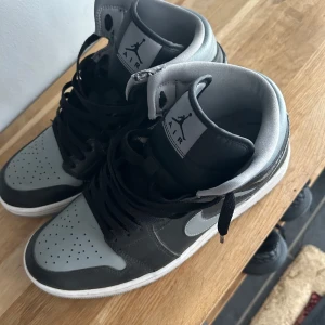 Nike Air Jordans i svart och grå - Säljer ett par Nike Air Jordans i svart och grå med högt skaft och klassisk vit sula. Skorna har snörning och den ikoniska swoosh-loggan på sidan. Perfekta för dig som gillar streetwear och vill ha en stilren sneaker.