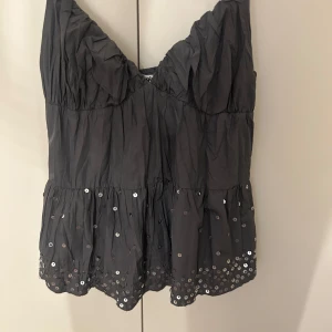 Svart paljettprydd linnetopp - Säljer en svart linnetopp med smala axelband och v-ringning. Toppen har glittriga paljetter längs nederkanten och en lätt rynkad struktur. Perfekt för dig som vill ha något extra till outfiten!Har en liten defekt syns på sista bilden men inget som märks