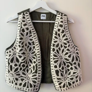 Olivgrön väst med virkad spets från Zara - Säljer en olivgrön väst från Zara med vit virkad spets i blommigt mönster över hela västen. Västen är ärmlös och har en öppen front, perfekt för att ge outfiten en bohemisk touch.