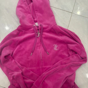 Rosa velour hoodie från Juicy Couture - Säljer en ikonisk rosa hoodie från Juicy Couture i mjuk velour. Jackan har dragkedja, huva, långa ärmar och glittriga Juicy-loggor både fram och bak. Perfekt för dig som vill ha en statement-jacka med lyxig känsla.