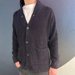 Mörkgrå stickad kofta med knappar - Säljer en mörkgrå stickad kofta med knappar framtill och två fickor. Koftan har lång ärm och en mjuk, strukturerad yta. Perfekt att slänga över en t-shirt för en avslappnad look.