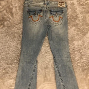 Ljusblå True Religion Joey jeans med slitningar - Säljer ett par ljusblå True Religion Joey jeans med låg midja och flare-ben. Jeansen har slitna detaljer på framsidan och ikoniska bakfickor med broderade sömmar i orange och gul. Klassisk knappstängning och tydlig True Religion-lapp bak.