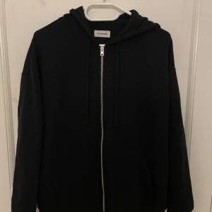 Svart hoodie med dragkedja från YOURTURN - Säljer en svart hoodie från YOURTURN med dragkedja framtill och huva. Tröjan har långa ärmar och en klassisk, enkel design som passar till det mesta. Perfekt för dig som gillar en stilren look.