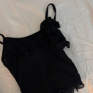 Svart linnetopp med volang och blomdetalj - Snygg svart linnetopp med tunna axelband och volangdetaljer längs ringningen. Toppen har en dekorativ blomma på ena axelbandet som ger en extra touch. Perfekt för dig som gillar stilrena men ändå unika plagg.