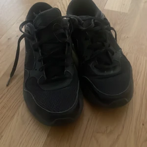 Svarta sneakers från Nike - Svarta sneakers från Nike med klassisk design och svarta snören. Skorna har rund tå och platt sula, vilket gör dem bekväma för vardagsbruk. Tillverkade i syntetmaterial med diskreta detaljer och meshpartier för extra andningsförmåga.