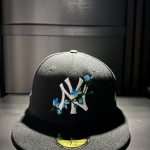 Svart New Era keps med broderade detaljer - Snygg svart keps från New Era med stor vit NY-logga framtill dekorerad med blå blommor. Baksidan har en broderad World Series 1996-logga i blått och vitt. Klassisk rak skärm och broderad New Era-logga på sidan.