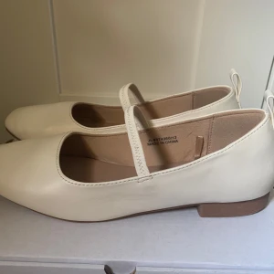 Vita ballerinaskor med rem från New Look - Säljer ett par vita ballerinaskor från New Look med klassisk rem över foten och låg klack. Skorna har rund tå och är perfekta för sommaren. 