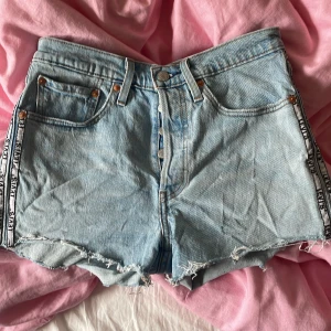 Levi's 501  ljusblå jeansshorts med fransar - Ljusblå jeansshorts från Levi's med hög midja och råa fransar nertill. Klassisk femficksmodell med knappgylf och dekorativa band med text längs sidorna. Perfekta för sommaren och har en avslappnad, trendig look.