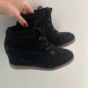 Svarta kilklackade sneakers med snörning - Säljer ett par svarta Stella marant liknade sneakers med kilklack och snörning. Skorna har en rund tå och platt gummisula i kontrasterande beige färg. Tillverkade i mocka för en mjuk och stilren look. 