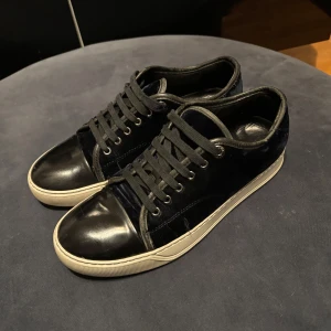 Svarta sneakers från Lanvin i mocka - Säljer ett par svarta sneakers från Lanvin med ovandel i mocka och detaljer i blankt skinn. Skorna har rund tå, snörning och vit platt sula. Klassisk och stilren design med lyxig känsla.