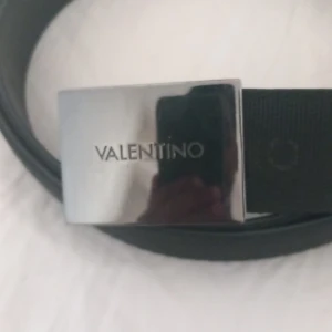 Svart bälte från Valentino med silverfärgat spänne - Snyggt svart bälte från Valentino med ett stilrent, rektangulärt spänne i silverfärg. Bältet har en diskret struktur i materialet och logotypen är ingraverad på spännet. Perfekt accessoar för att lyfta din outfit.