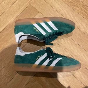 Adidas Gazelle gröna sneakers - Säljer ett par gröna Adidas Gazelle sneakers med klassiska vita ränder och vit häl. Storlek 42 2/3 (passar 42) ny pris ca 1400. Använda men i gott skick