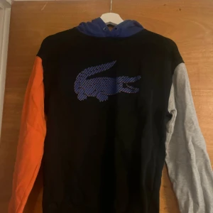 Flerfärgad hoodie från Lacoste - hoodie från Lacoste med blå huva, svart framsida, orange ärm och grå ärm. Stor blå Lacoste-logga på bröstet.  Limited edition hoodie