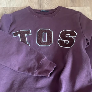 Vinröd sweatshirt från Tiger of Sweden - Säljer en vinröd sweatshirt från Tiger of Sweden med stora bokstäver 'TOS' framtill. Tröjan har rund halsringning och långa ärmar. Perfekt för en avslappnad stil.