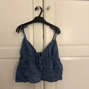 Blå croppad linnetopp med knyt - Säljer en blå croppad linnetopp med smala axelband och knyt framtill. Toppen har volangdetaljer och är perfekt för varma dagar. Snygg och enkel att matcha till olika outfits.