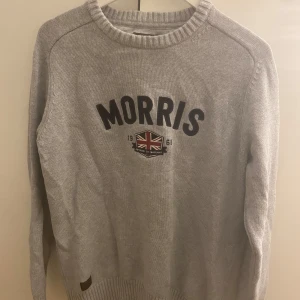 Grå stickad tröja från Morris - Säljer en grå stickad tröja från Morris med rund halsringning och svart textlogga samt brittisk flagga framtill. Tröjan har långa ärmar och ribbade muddar vid ärmslut och nederkant. Perfekt för kyligare dagar. Passar både L/M