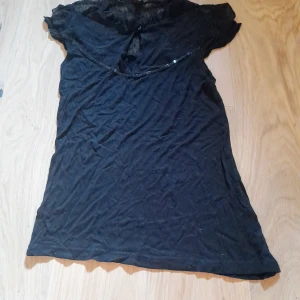 Svart blus med spetsdetaljer från Butik - Svart blus från Butik med spets upptill och korta ärmar. Blusen har en liten knapp i nacken och dekorativt band framtill. Perfekt för en stilren look med extra detalj. Paljetter som glittrar. Använd enstaka gång. Den passar en storlek xs-small beroende på hur tight en vill ha den