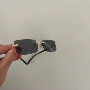 Svarta rektangulära solglasögon med gulddetaljer - Stilrena solglasögon med rektangulära svarta glas och tunna guldfärgade bågar. Glasögonen har minimalistisk design utan synlig ram runt glasen och svarta skalmar. Perfekta för en trendig look.