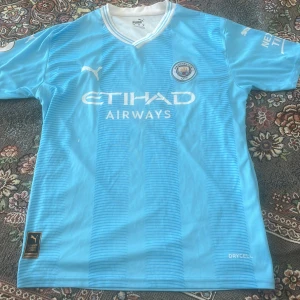 Manchester City fotbollströja Haaland 9 - Säljer en ljusblå Manchester City fotbollströja från Puma med vitt tryck, Etihad Airways sponsorlogga och klubbmärke på bröstet. Nummer 9 och Haaland på ryggen. Klassisk V-ringning och korta ärmar. Materialet är lätt och andas.