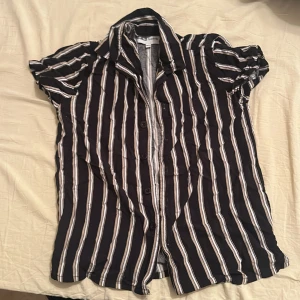 Svart och vit randig kortärmad skjorta - Kortärmad skjorta med svarta och vita ränder samt inslag av beige. Skjortan har klassisk krage och knäppning framtill. Perfekt för en avslappnad och stilren look.