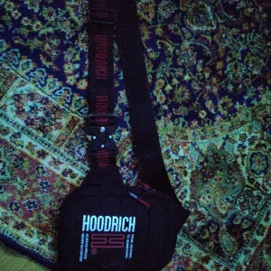 Svart axelväska från Hoodrich - Säljer en svart axelväska från Hoodrich med justerbart axelband och vit/röd logga framtill. Väskan har dragkedja och tryckt text på framsidan. Perfekt för att bära det viktigaste när du är på språng.