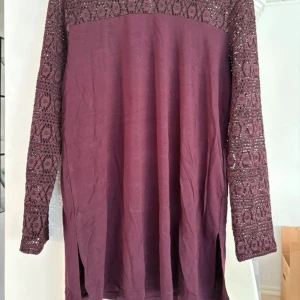 Plommonlila klänning/tunika med spetsdetaljer - Plommonlila långärmad tunika/klänning från Flash Woman med vackra virkade spetsdetaljer upptill och på ärmarna. Modellen är rak med små slitsar i sidorna och har en mjuk, luftig känsla. Oanvänd med prislapp