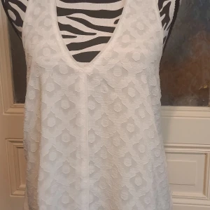Vit ärmlös topp från Guess - Säljer en vit ärmlös topp från Guess i storlek M. Toppen har ett diskret geometriskt mönster och v-ringning. Perfekt för varma dagar och enkel att matcha med olika outfits.