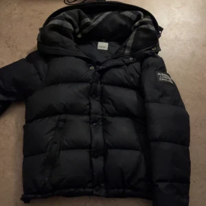 Svart dunjacka från Burberry - Snygg svart dunjacka från Burberry med huva och vadderad design. Jackan har en patch på ärmen och klassisk Burberry-etikett på insidan. Perfekt för kyliga dagar och har en sportig, modern look.