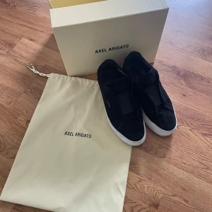 Svarta sneakers från Axel Arigato - Säljer ett par svarta sneakers från Axel Arigato med vit sula och diskret logga på sidan. Skorna är i mocka och har en stilren design utan snörning, med elastiskt band över vristen. Kommer med originalpåse och låda.