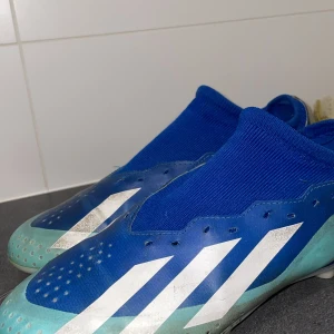 Blåa Adidas fotbollsskor - Säljer ett par blåa Adidas fotbollsskor med vita ränder och turkosa detaljer. Skorna har en strumpliknande passform utan snörning och är designade för spel på konstgräs eller gräs. Perfekta för dig som vill ha bra grepp och stil på planen.