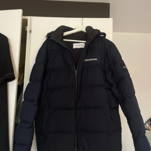 Mörkblå pufferjacka från Calvin Klein Jeans - Säljer en mörkblå pufferjacka från Calvin Klein Jeans med huva och dragkedja. Jackan har logotyp på bröstet och ärmen, samt en klassisk quiltad design som håller värmen. Perfekt för kyliga dagar.   Gå gärna in på Vinted och köp, mycket smidigare leverans och betalningsmetod! - sessaskogen där