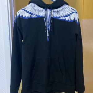 Marcelo Burlon hoodie - Säljer en svart hoodie från Marcelo Burlon med unikt blått och vitt vingmönster över axlar och huva. Tröjan har lång ärm, huva med dragsko och en stor magficka. Perfekt för dig som vill sticka ut.